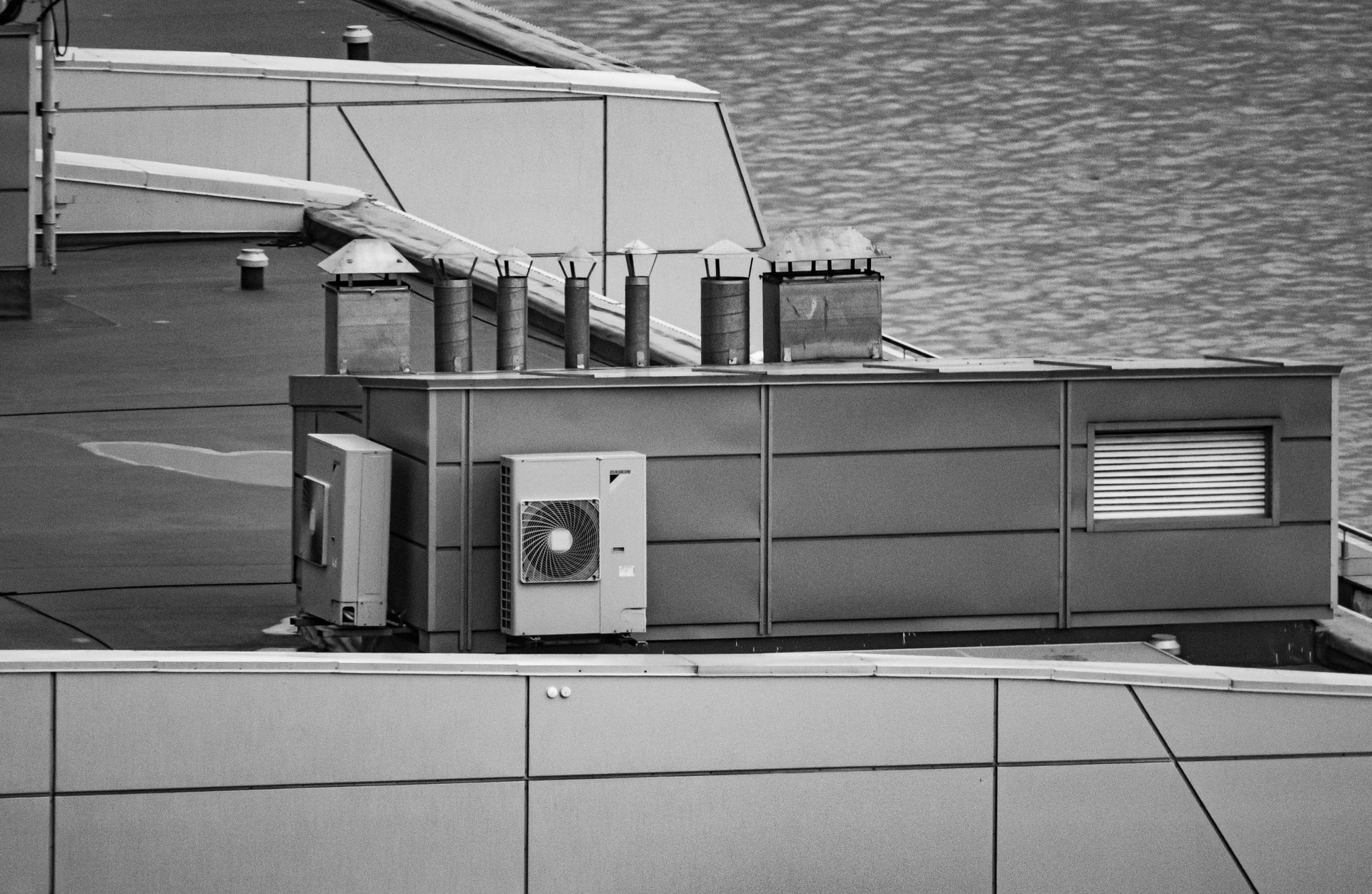Rooftop HVAC equipment with vents and a wall-mounted unit on a gray building, water visible in the background.